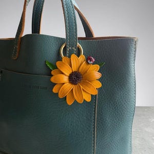 May include: Teal leather tote bag with a sunflower and ladybug charm. The bag has a structured design with double handles and a small pocket. The sunflower charm is orange and brown, with green leaves and a red ladybug.