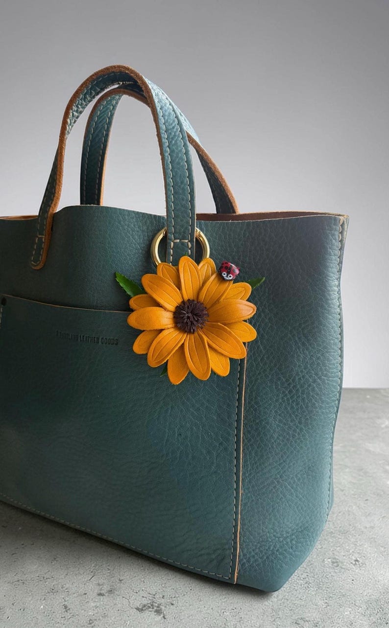 May include: Teal leather tote bag with a sunflower and ladybug charm. The bag has a structured design with double handles and a small pocket. The sunflower charm is orange and brown, with green leaves and a red ladybug.