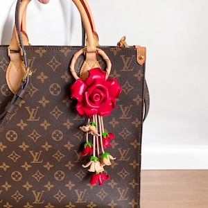 May include: A brown leather handbag with a red rose and pink flower charm. The bag has a brown and tan monogram pattern.