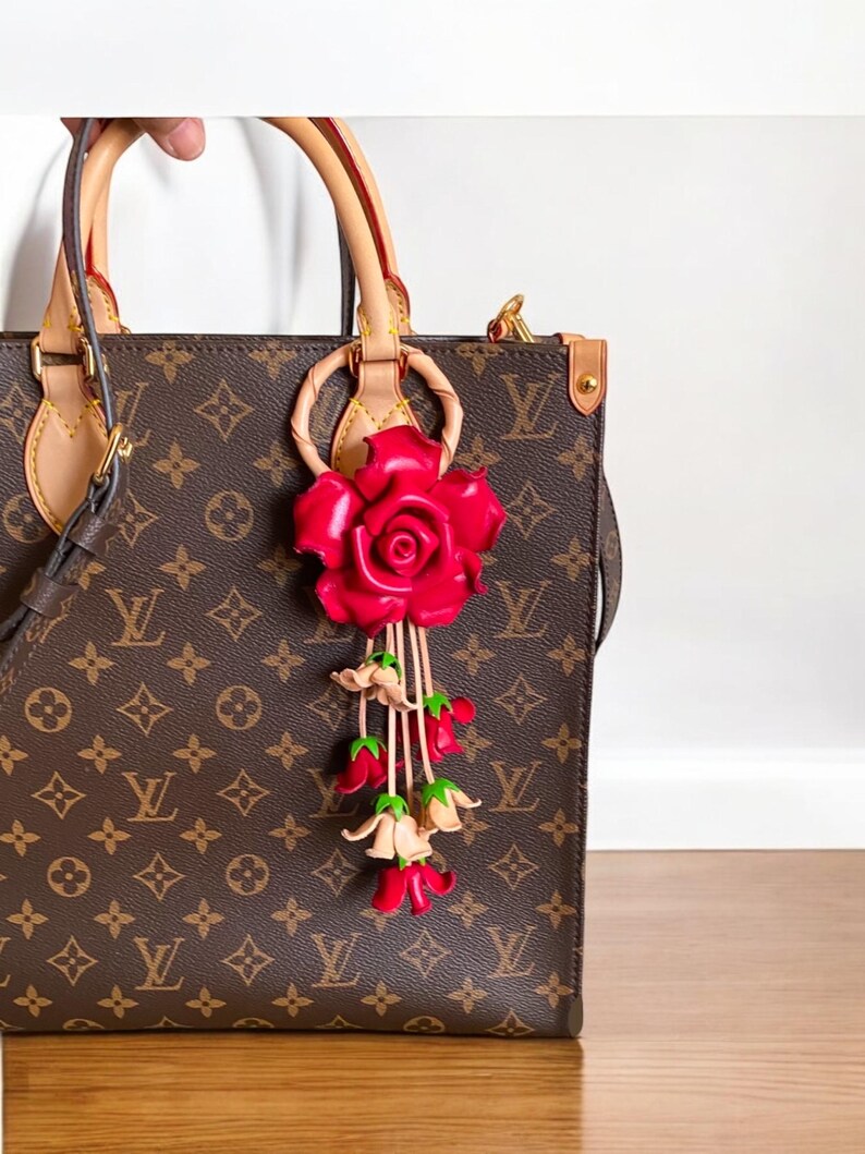 May include: A brown leather handbag with a red rose and pink flower charm. The bag has a brown and tan monogram pattern.