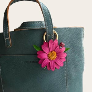 May include: A teal leather tote bag with a pink flower and ladybug charm. The bag has a top handle and a gold ring. The flower has a yellow center and green leaves. The ladybug is red with black spots.