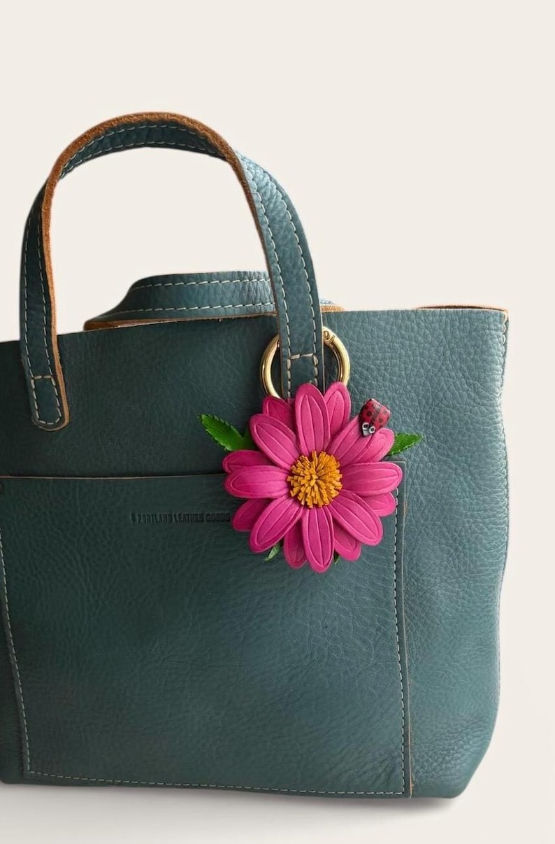 May include: A teal leather tote bag with a pink flower and ladybug charm. The bag has a top handle and a gold ring. The flower has a yellow center and green leaves. The ladybug is red with black spots.