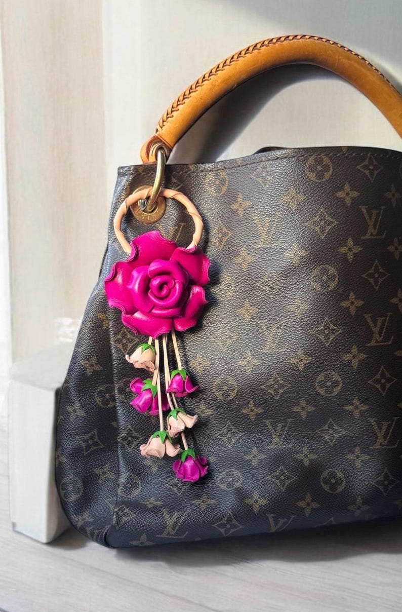 May include: A brown leather handbag with a pink leather rose charm. The rose charm has a pink leather flower with a green stem and leaves. The handbag has a brown leather strap and a brown and tan monogram pattern.