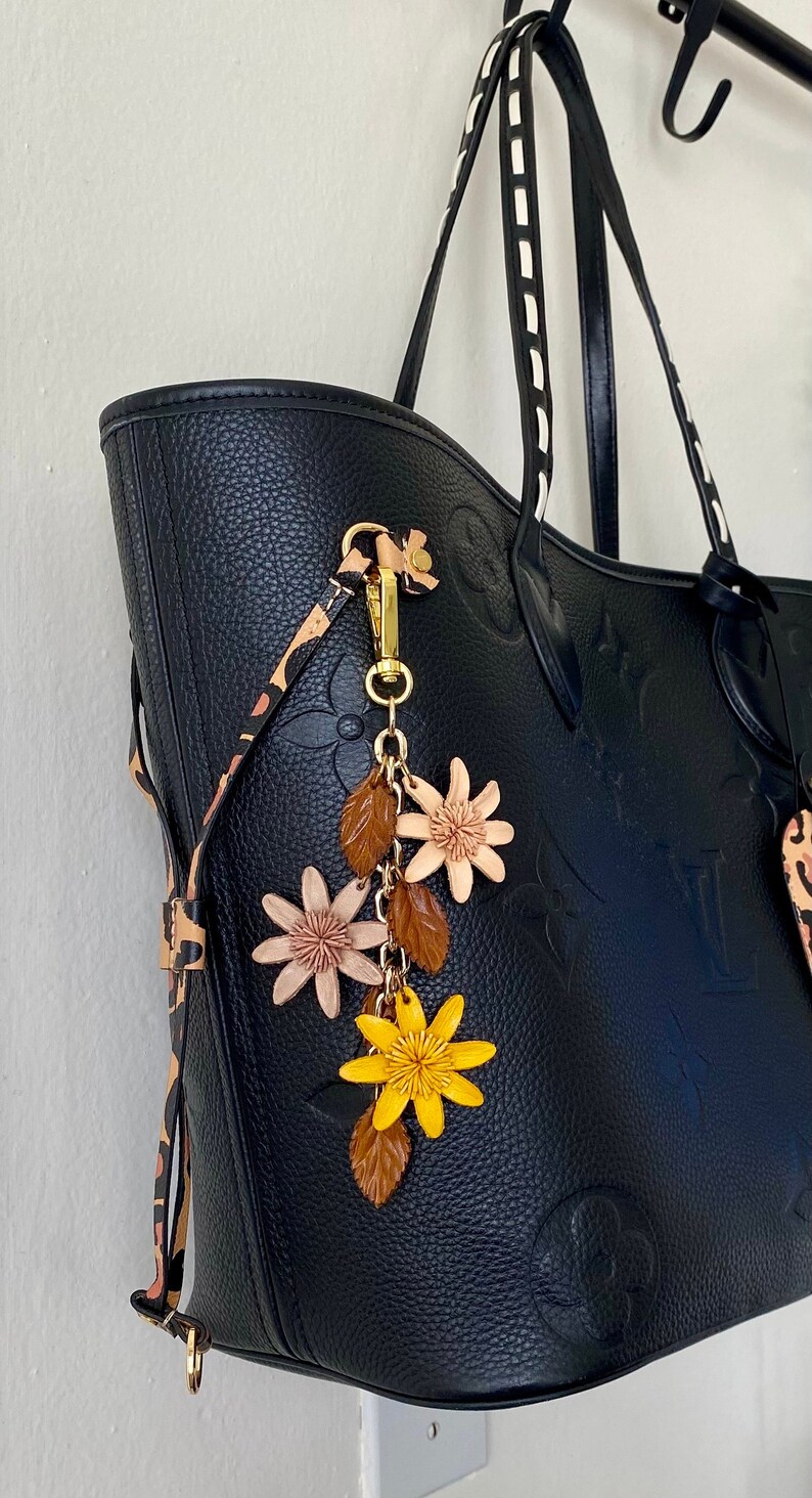 K&ouml;nnte beinhalten: Schwarze Tragetasche mit gepr&auml;gtem Blumenmuster, mit einem dekorativen Anh&auml;nger. Der Anh&auml;nger enth&auml;lt einen goldfarbenen Verschluss, braune Bl&auml;tter und rosa und gelbe Blumenakzente. Die Tasche hat einen Leopardenmuster-Gurt.