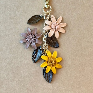 K&ouml;nnte beinhalten: Ein floraler Schl&uuml;sselanh&auml;nger mit einem goldfarbenen Verschluss. Der Schl&uuml;sselanh&auml;nger zeigt drei Lederblumen in Rosa- und Gelbt&ouml;nen mit dunkelgr&uuml;nen Bl&auml;ttern. Der Text "NATURALLY MUTED" steht oben.