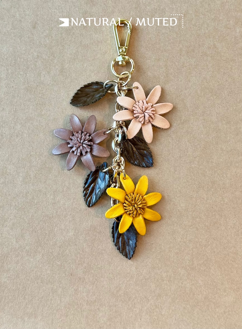 K&ouml;nnte beinhalten: Ein floraler Schl&uuml;sselanh&auml;nger mit einem goldfarbenen Verschluss. Der Schl&uuml;sselanh&auml;nger zeigt drei Lederblumen in Rosa- und Gelbt&ouml;nen mit dunkelgr&uuml;nen Bl&auml;ttern. Der Text "NATURALLY MUTED" steht oben.