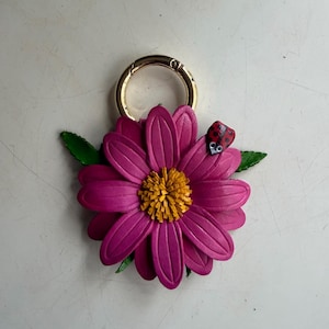 May include: A magenta leather flower keychain with a gold-colored ring. The flower has a yellow center and green leaves. A small red ladybug with black spots rests on a petal.
