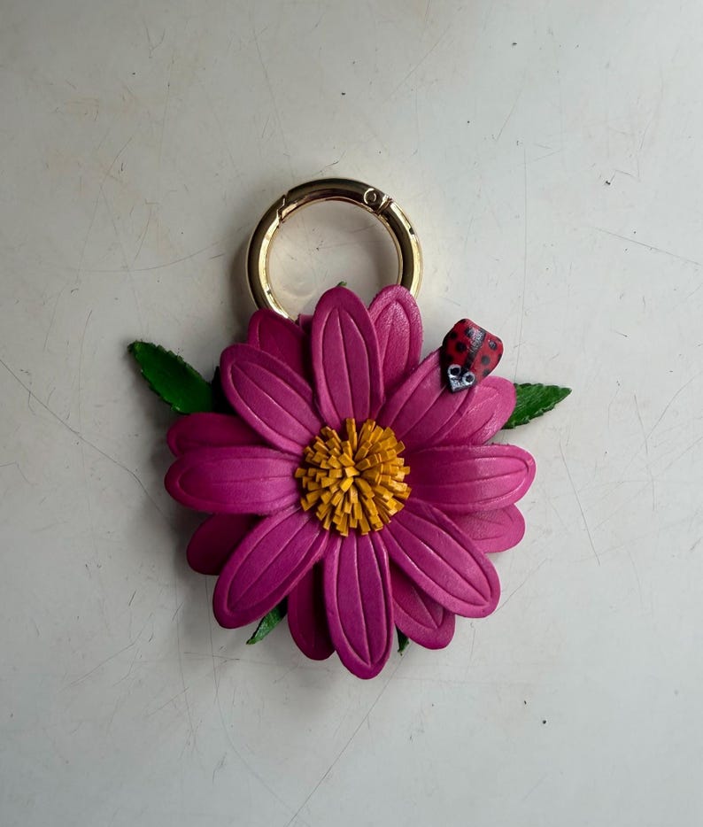 May include: A magenta leather flower keychain with a gold-colored ring. The flower has a yellow center and green leaves. A small red ladybug with black spots rests on a petal.