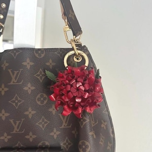 Op de afbeelding: Een rode leren bloemhanger met groene bladeren hangt aan een goudkleurige sluiting die aan een bruine designertas is bevestigd. De handtas heeft een herhalend patroon van bruine logo's.