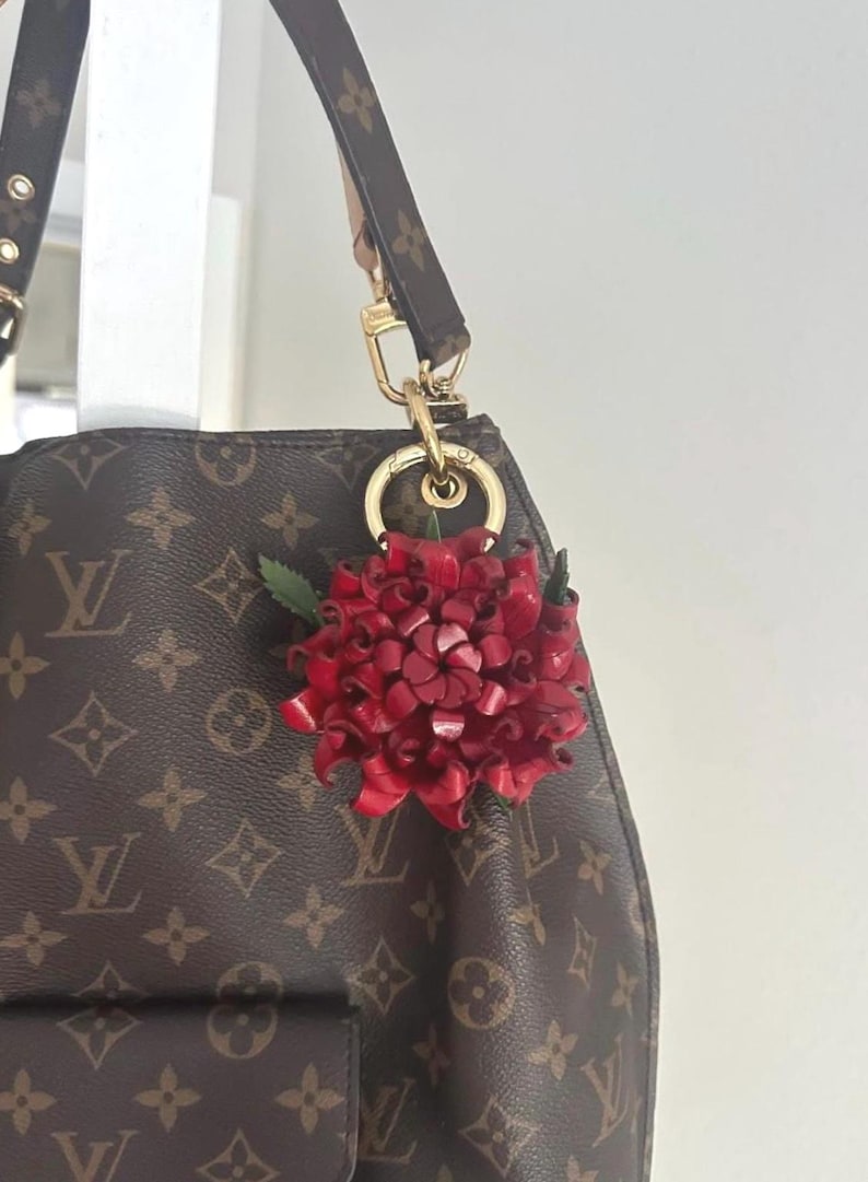 Op de afbeelding: Een rode leren bloemhanger met groene bladeren hangt aan een goudkleurige sluiting die aan een bruine designertas is bevestigd. De handtas heeft een herhalend patroon van bruine logo's.