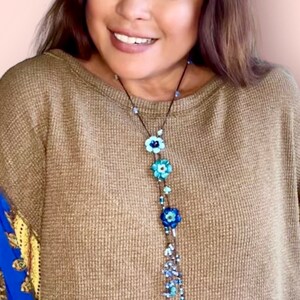 May include: A long necklace featuring a dark cord adorned with three distinct blue flower-shaped beads, interspersed with smaller light blue and dark blue glass beads, and finished with a dangling cluster of various blue beads at the bottom.