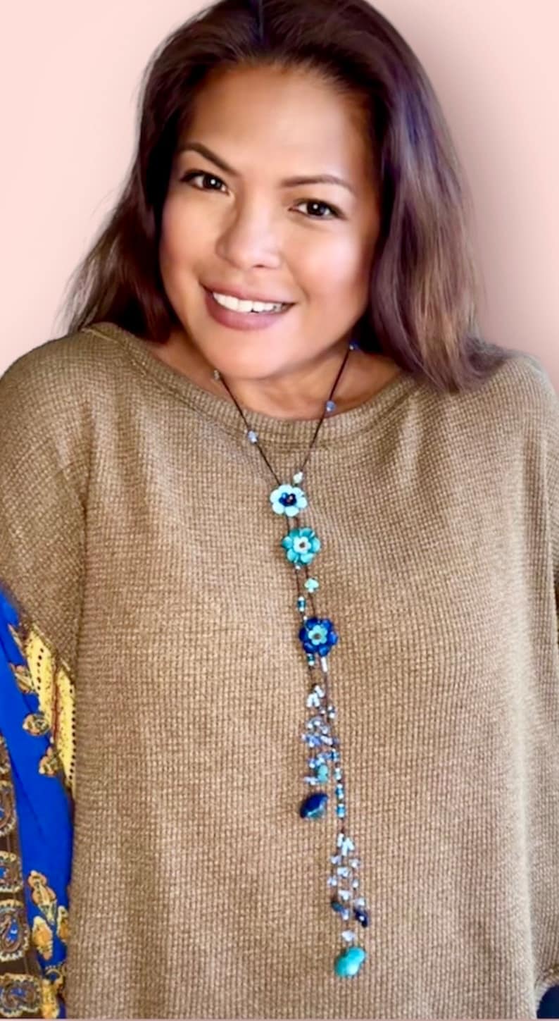 May include: A long necklace featuring a dark cord adorned with three distinct blue flower-shaped beads, interspersed with smaller light blue and dark blue glass beads, and finished with a dangling cluster of various blue beads at the bottom.