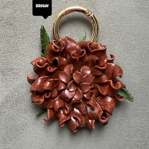 Op de afbeelding: Een bruine leren bloem sleutelhanger met een goudkleurige ring. De bloem heeft gedetailleerde, gelaagde bloemblaadjes en kleine groene blaadjes. Het woord "BROWN" staat in een zwart kader boven de sleutelhanger.
