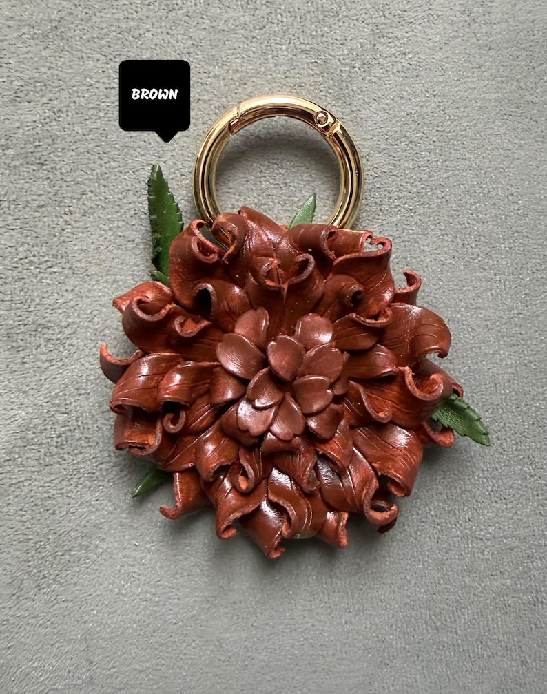 Op de afbeelding: Een bruine leren bloem sleutelhanger met een goudkleurige ring. De bloem heeft gedetailleerde, gelaagde bloemblaadjes en kleine groene blaadjes. Het woord "BROWN" staat in een zwart kader boven de sleutelhanger.