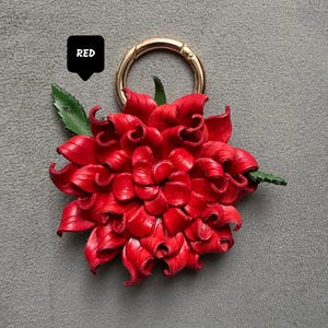 Op de afbeelding: Een felrode leren bloem met een goudkleurige sluiting. De bloem heeft gedetailleerde bloemblaadjes en een groen blad. Het woord "RED" staat in een zwarte tekstballon.