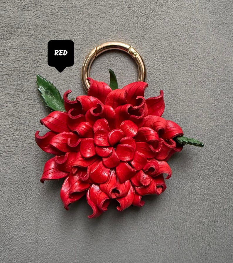 Op de afbeelding: Een felrode leren bloem met een goudkleurige sluiting. De bloem heeft gedetailleerde bloemblaadjes en een groen blad. Het woord "RED" staat in een zwarte tekstballon.