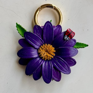 May include: A purple leather flower keychain with a gold-colored clasp. The flower has a yellow center and green leaves. A small red ladybug with black spots is perched on a petal.