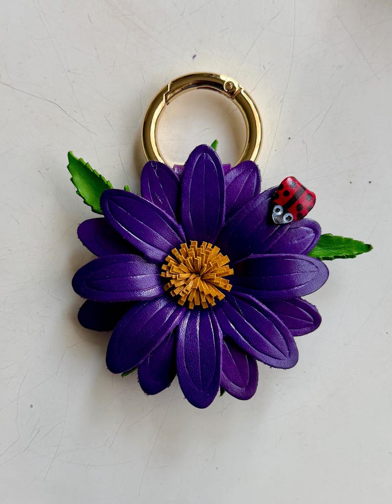 May include: A purple leather flower keychain with a gold-colored clasp. The flower has a yellow center and green leaves. A small red ladybug with black spots is perched on a petal.