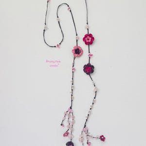 May include: A long necklace featuring a black cord adorned with pink and dark purple flower-shaped beads, interspersed with small clear and light pink crystal-like beads. The text "Pretty Pink combo" is printed in pink near the center of the cord.