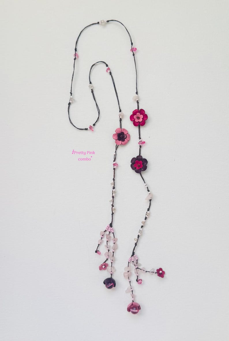 May include: A long necklace featuring a black cord adorned with pink and dark purple flower-shaped beads, interspersed with small clear and light pink crystal-like beads. The text "Pretty Pink combo" is printed in pink near the center of the cord.