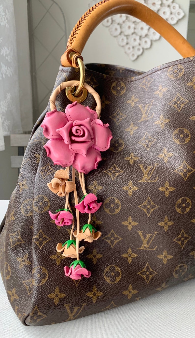 May include: A pink leather rose charm with a dangling chain of smaller pink and beige roses. The charm is attached to a brown leather handbag with a monogram pattern.