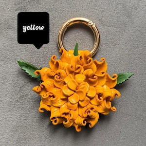 Op de afbeelding: Een gele leren bloem met groene bladeren en een goudkleurige metalen ring. Het woord "yellow" staat in een zwarte tekstballon. De bloem heeft gedetailleerde gekrulde bloemblaadjes.