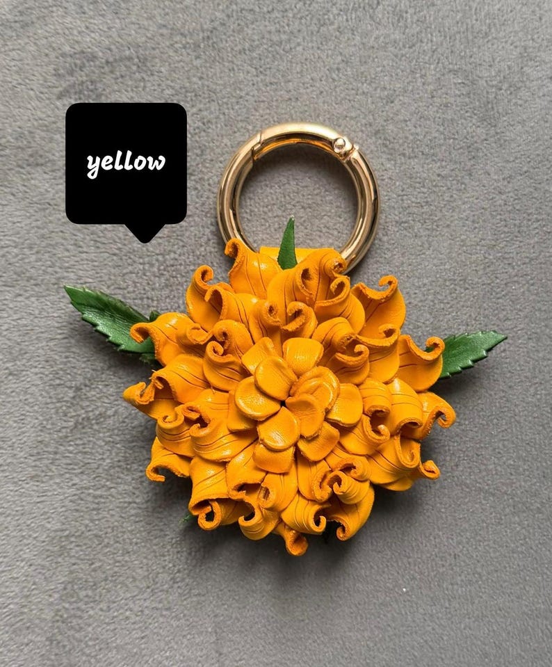 Op de afbeelding: Een gele leren bloem met groene bladeren en een goudkleurige metalen ring. Het woord "yellow" staat in een zwarte tekstballon. De bloem heeft gedetailleerde gekrulde bloemblaadjes.