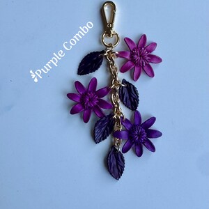 K&ouml;nnte beinhalten: Ein lila Blumen-Schl&uuml;sselanh&auml;nger mit einem goldfarbenen Verschluss und einer Kette. Der Schl&uuml;sselanh&auml;nger hat drei lila Blumen und dunkelviolette Bl&auml;tter. Der Text "Purple Combo" befindet sich in der oberen linken Ecke.