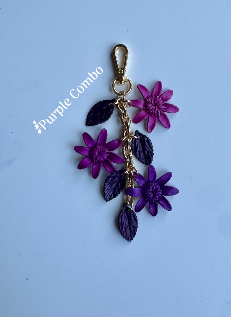 K&ouml;nnte beinhalten: Ein lila Blumen-Schl&uuml;sselanh&auml;nger mit einem goldfarbenen Verschluss und einer Kette. Der Schl&uuml;sselanh&auml;nger hat drei lila Blumen und dunkelviolette Bl&auml;tter. Der Text "Purple Combo" befindet sich in der oberen linken Ecke.