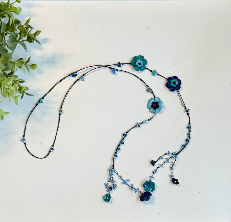 May include: A long necklace featuring a dark cord strung with various shades of blue and turquoise flower-shaped beads, interspersed with small, irregular light blue and dark blue stone chips, ending in a dangling floral arrangement.