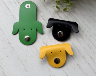 May include: Three leather cord organisers shaped like dog heads in green, black, and yellow. Each organiser features two small punched holes for eyes and a silver-toned metal snap button closure at the bottom to secure cords in place.