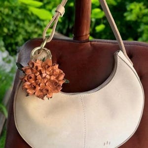 Op de afbeelding: Cr&egrave;mekleurige, halvemaanvormige handtas met bijpassende riem. Een decoratieve leren bloemhanger in bruin- en groentinten hangt aan de handgreep. De tas is gemaakt van zacht materiaal en heeft een ritssluiting.