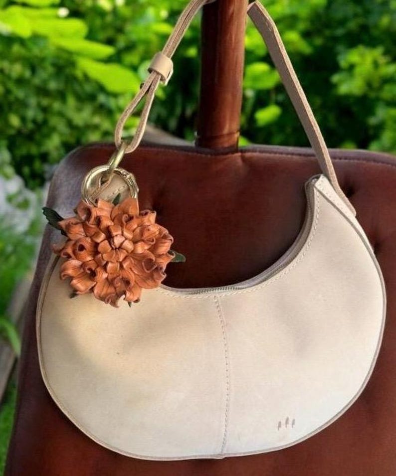 Op de afbeelding: Cr&egrave;mekleurige, halvemaanvormige handtas met bijpassende riem. Een decoratieve leren bloemhanger in bruin- en groentinten hangt aan de handgreep. De tas is gemaakt van zacht materiaal en heeft een ritssluiting.