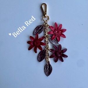 K&ouml;nnte beinhalten: Ein floraler Schl&uuml;sselanh&auml;nger mit einem goldfarbenen Verschluss und einer Kette. Der Schl&uuml;sselanh&auml;nger zeigt mehrere tiefrote Bl&uuml;ten und dunkelgraue Bl&auml;tter. Der Text "Bella Red" befindet sich in der oberen linken Ecke.