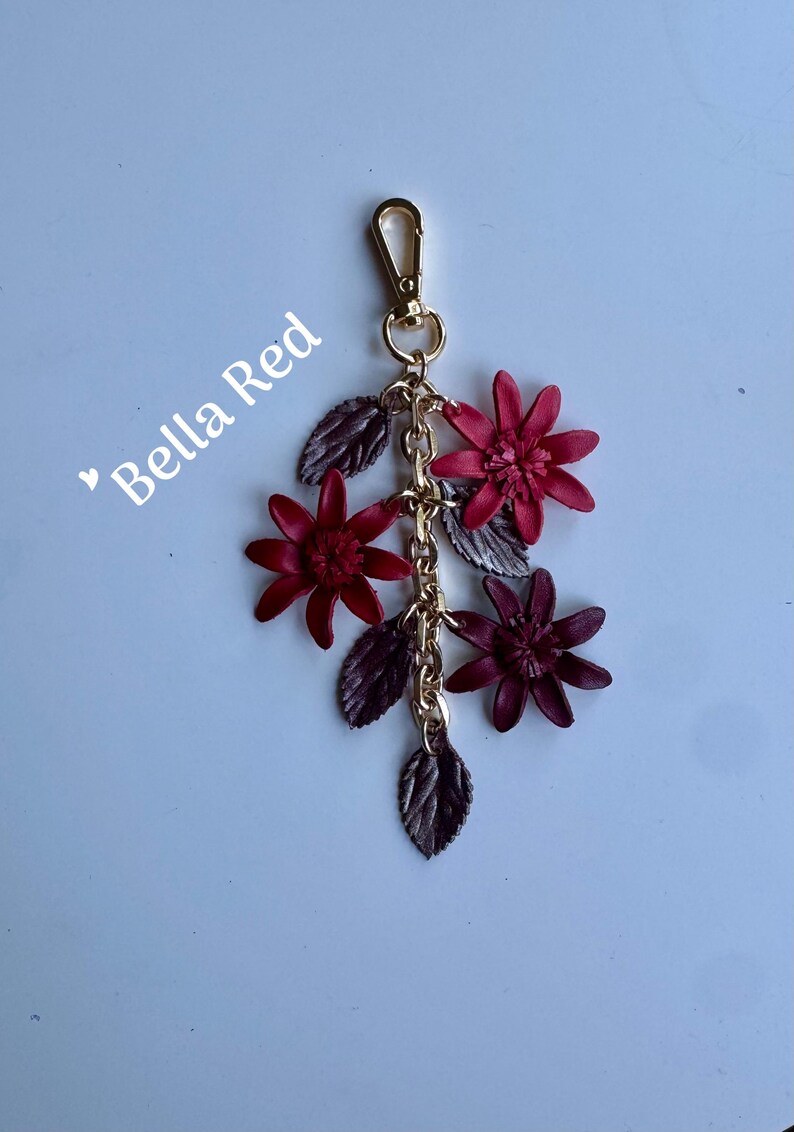 K&ouml;nnte beinhalten: Ein floraler Schl&uuml;sselanh&auml;nger mit einem goldfarbenen Verschluss und einer Kette. Der Schl&uuml;sselanh&auml;nger zeigt mehrere tiefrote Bl&uuml;ten und dunkelgraue Bl&auml;tter. Der Text "Bella Red" befindet sich in der oberen linken Ecke.