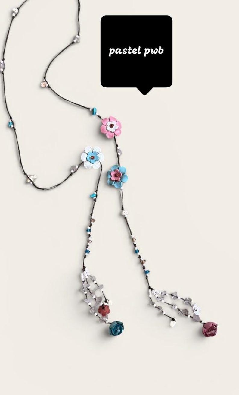 May include: A long necklace featuring a thin black cord adorned with small colorful beads and several layered leather-style flower charms in shades of pink, blue, and white. The ends feature dangling bead clusters and a black speech bubble with the text pastel pwb.