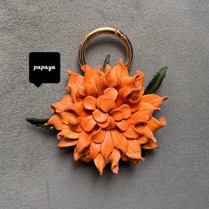 Op de afbeelding: Oranje leren bloemen sleutelhanger met een goudkleurige sluiting en groene bladeren. De bloem is gemaakt van gelaagde bloemblaadjes. Het woord "papaya" staat in een zwarte tekstballon. Decoratief accessoire.