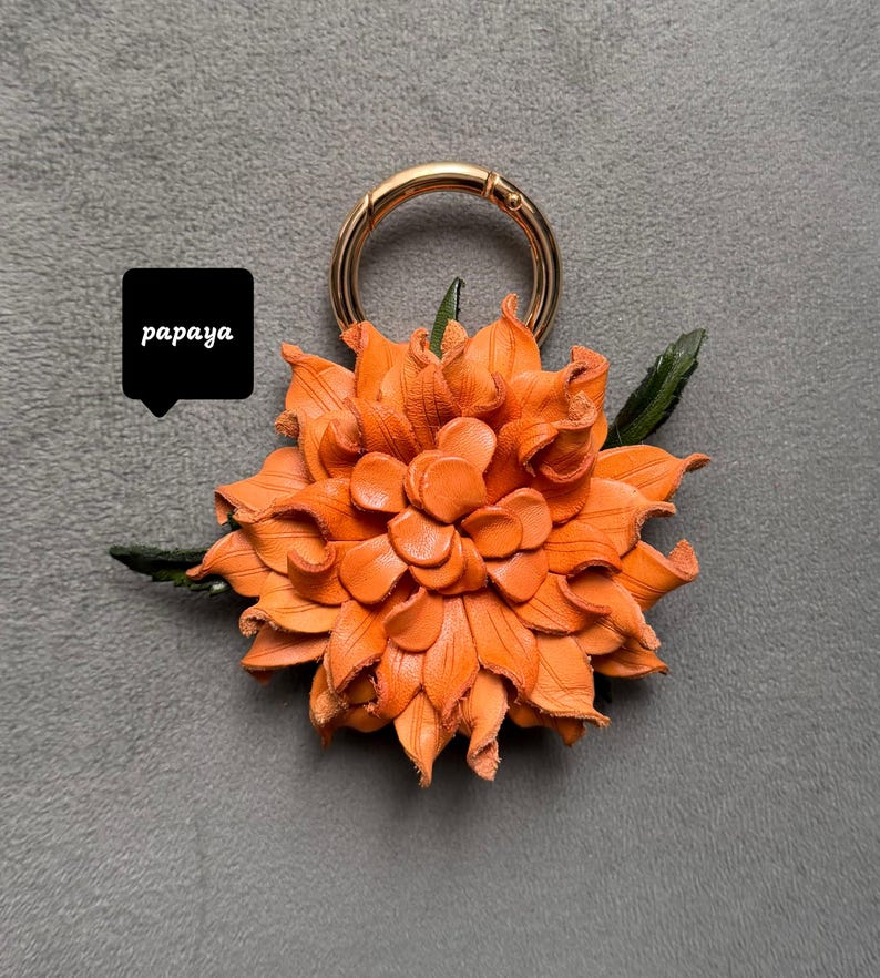 Op de afbeelding: Oranje leren bloemen sleutelhanger met een goudkleurige sluiting en groene bladeren. De bloem is gemaakt van gelaagde bloemblaadjes. Het woord "papaya" staat in een zwarte tekstballon. Decoratief accessoire.