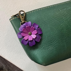 以下が含まれることがあります： 紫とピンクの色合いの革製の花のキーホルダー。真鍮の留め金が付いた緑色の革製ポーチに取り付けられています。花は複数の花びらの層と中央のボタンがあります。ポーチには白いステッチが施されています。