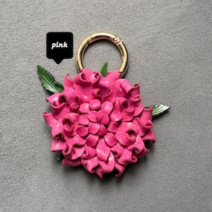 Op de afbeelding: Een felroze bloemvormige sleutelhanger met een goudkleurige ring. De bloem heeft een gedetailleerd bloemblaadjesontwerp en groene bladeren. Het woord "pink" staat in een zwarte tekstballon.