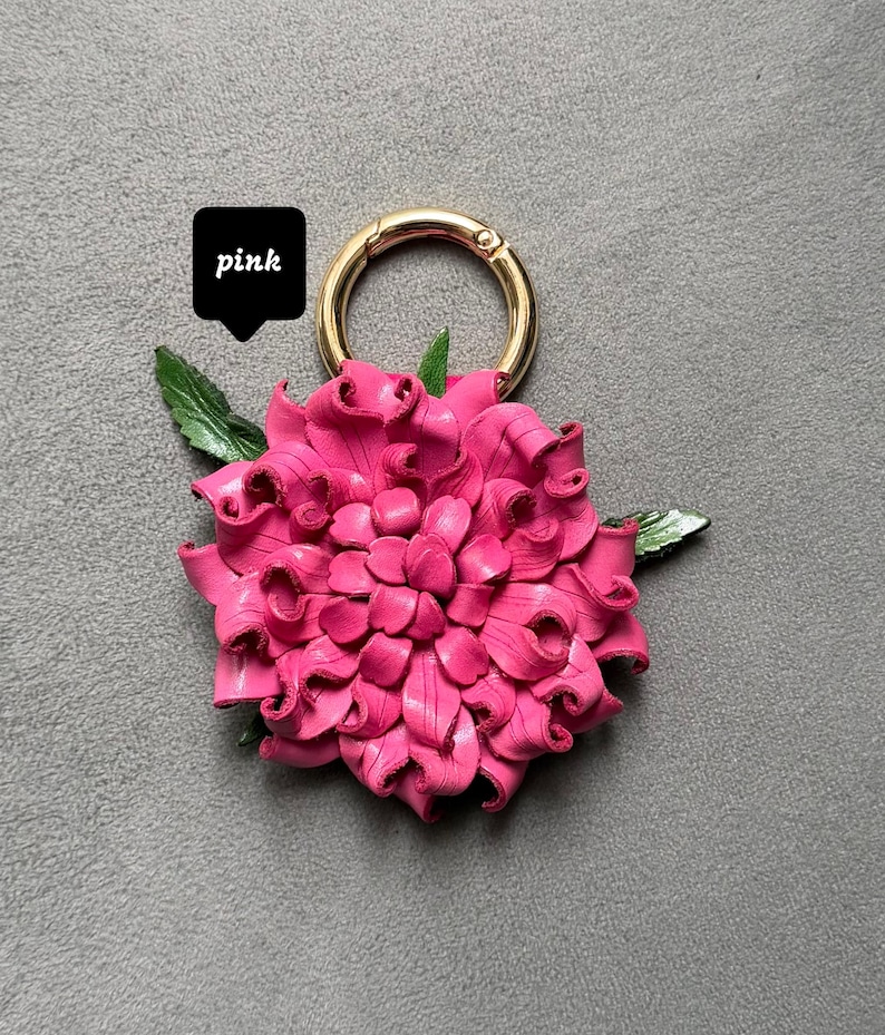 Op de afbeelding: Een felroze bloemvormige sleutelhanger met een goudkleurige ring. De bloem heeft een gedetailleerd bloemblaadjesontwerp en groene bladeren. Het woord "pink" staat in een zwarte tekstballon.