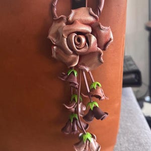 May include: A brown leather tote bag with a decorative rose and flower charm. The charm features a large rose and cascading bell-shaped flowers in shades of brown and green. The bag has a dark brown handle.