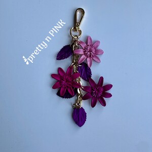 K&ouml;nnte beinhalten: Ein floraler Schl&uuml;sselanh&auml;nger mit einem goldfarbenen Verschluss und einer Kette. Der Schl&uuml;sselanh&auml;nger zeigt drei Bl&uuml;ten in Rosa- und Lilat&ouml;nen sowie lila Bl&auml;tter. Der Text "pretty n PINK" befindet sich in der oberen linken Ecke.