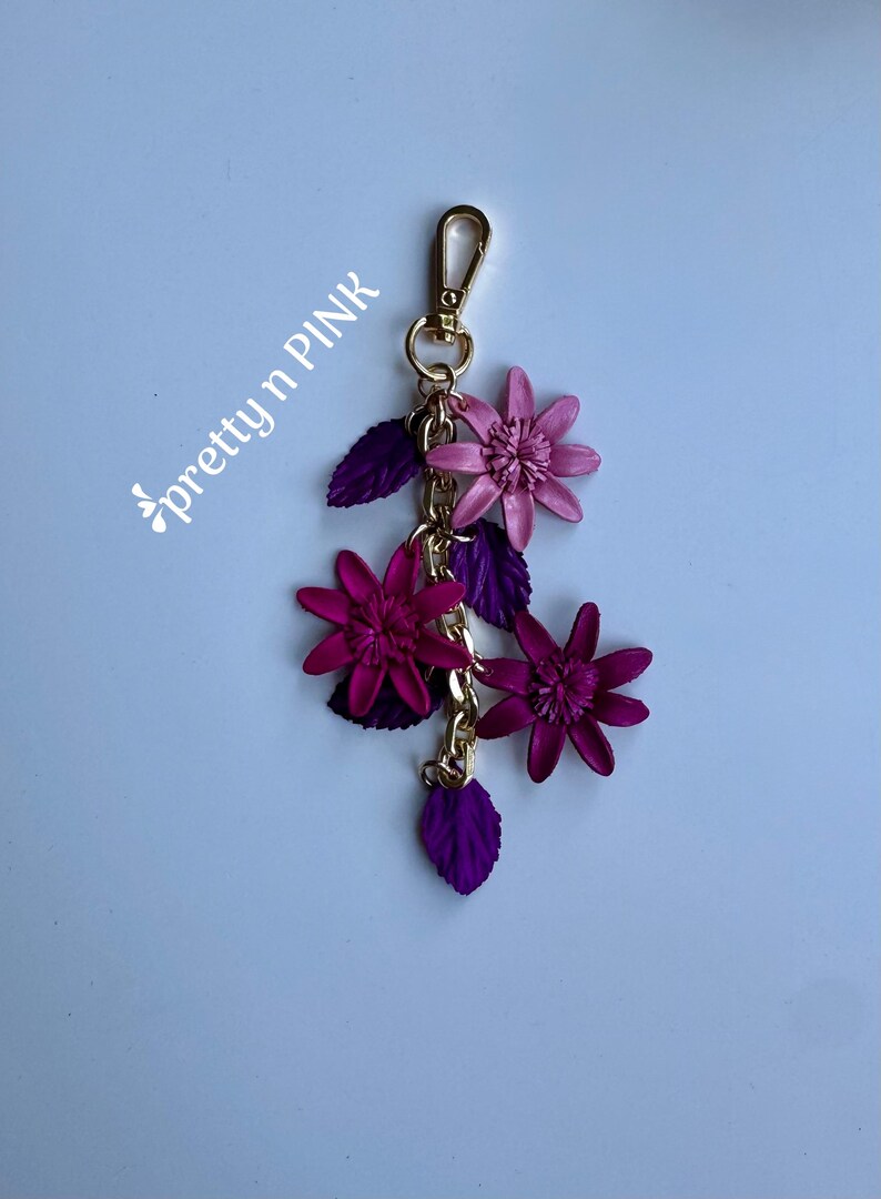 K&ouml;nnte beinhalten: Ein floraler Schl&uuml;sselanh&auml;nger mit einem goldfarbenen Verschluss und einer Kette. Der Schl&uuml;sselanh&auml;nger zeigt drei Bl&uuml;ten in Rosa- und Lilat&ouml;nen sowie lila Bl&auml;tter. Der Text "pretty n PINK" befindet sich in der oberen linken Ecke.