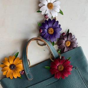 May include: A teal leather tote bag with gold hardware, adorned with colorful leather flower charms. The flowers include white, purple, brown, red, and yellow, each with a ladybug accent. The bag has a handle and a label that reads "Portland Leather Goods."