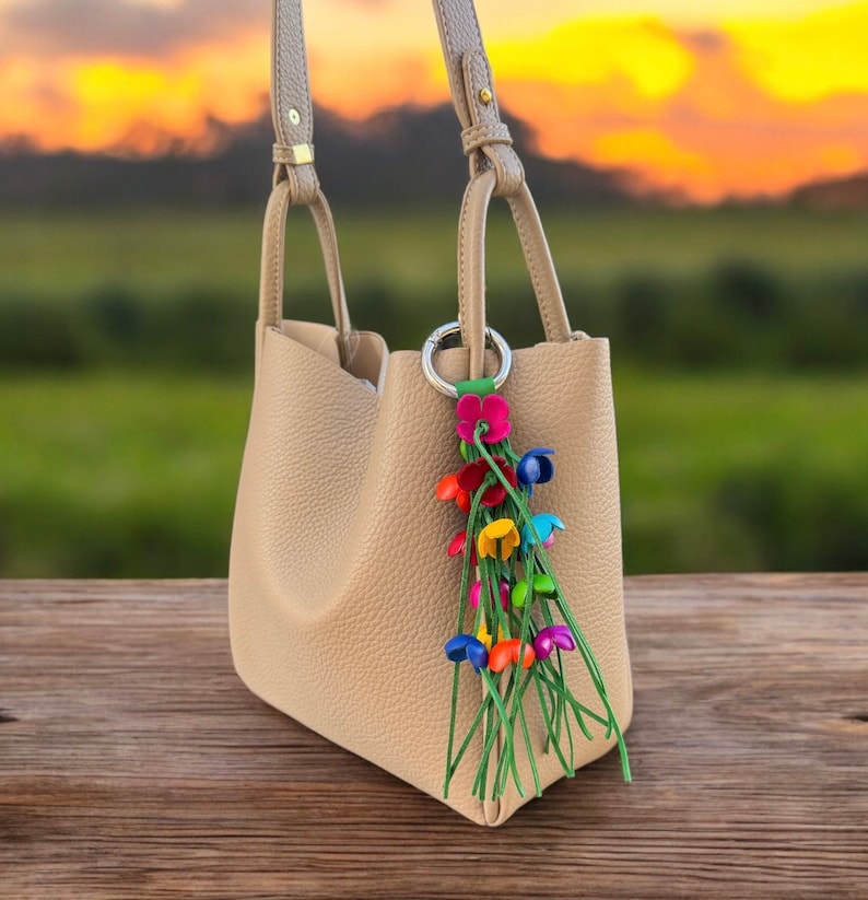 K&ouml;nnte beinhalten: Eine beige Lederhandtasche mit einem bunten Blumenanh&auml;nger an der Henkeltasche. Der Anh&auml;nger besteht aus einer Gruppe kleiner, bunter Blumen mit gr&uuml;nen Stielen.