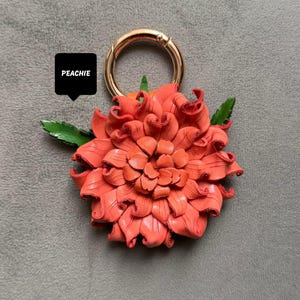 Op de afbeelding: Een koraalkleurige leren bloem met een goudkleurige ringsluiting. De bloem heeft gedetailleerde bloemblaadjes en groene bladeren. Het woord "PEACHIE" staat in een zwarte tekstballon.