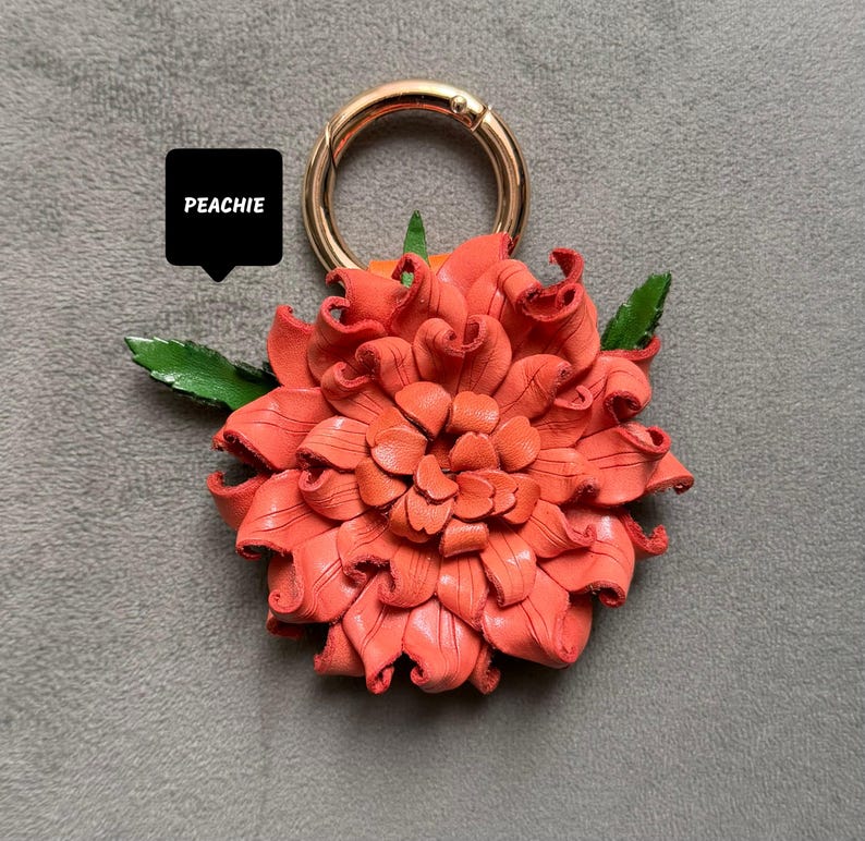 Op de afbeelding: Een koraalkleurige leren bloem met een goudkleurige ringsluiting. De bloem heeft gedetailleerde bloemblaadjes en groene bladeren. Het woord "PEACHIE" staat in een zwarte tekstballon.