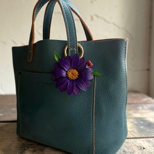 May include: Teal leather tote bag with a front pocket and brown leather handles. A large purple flower with a yellow center and a small ladybug charm are attached to the bag. The bag has gold-colored hardware.