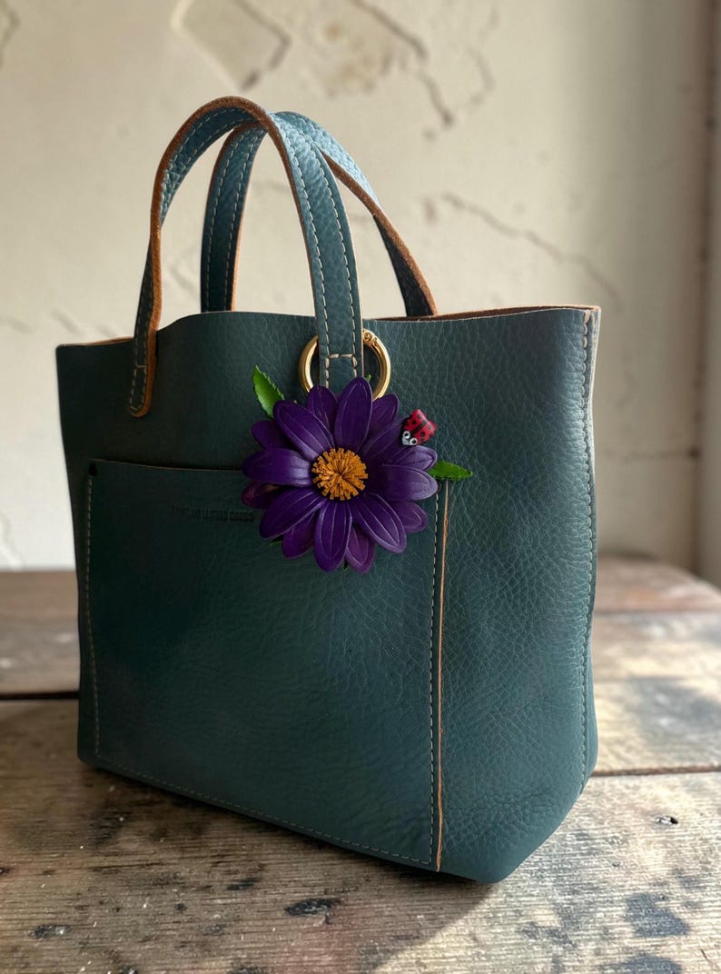 May include: Teal leather tote bag with a front pocket and brown leather handles. A large purple flower with a yellow center and a small ladybug charm are attached to the bag. The bag has gold-colored hardware.